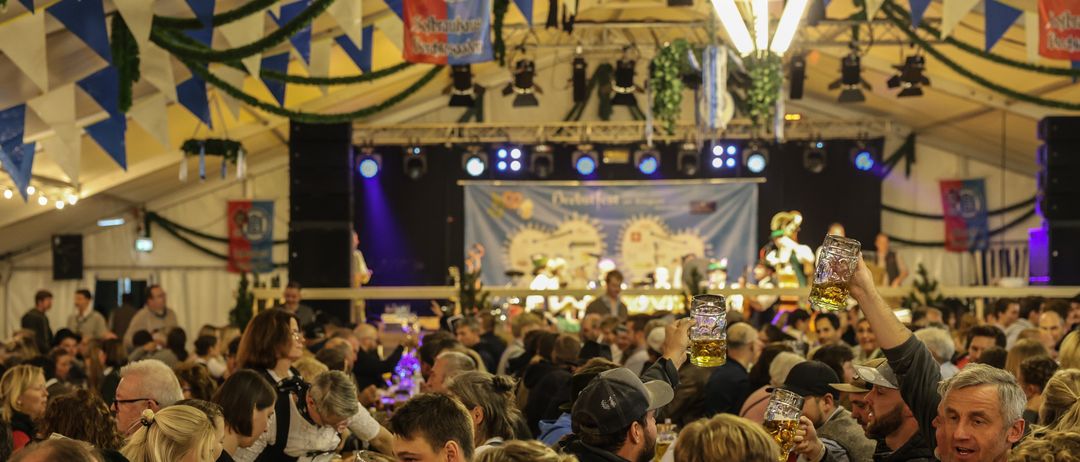 Eine große Menschenmenge genießt Bier bei einer festlichen Veranstaltung, mit Bannern und Flaggen, die das Zelt schmücken. Eine Bühne mit einem Banner und Scheinwerfern im Hintergrund, während die Menschen Biergläser halten.