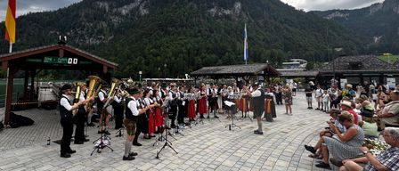 Eine Blaskapelle spielt im Freien auf einem gepflasterten Bereich, mit Bergen und einem Fahnenmast im Hintergrund. Das Publikum sitzt an der Seite.