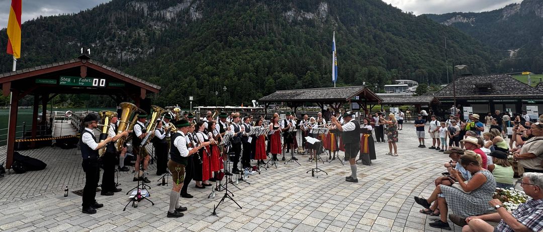 Eine Blaskapelle spielt im Freien auf einem gepflasterten Bereich, mit Bergen und einem Fahnenmast im Hintergrund. Das Publikum sitzt an der Seite.