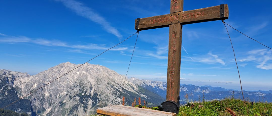 Ein Holzkreuz steht auf einem Berggipfel, mit einem kleinen Holztisch darunter. Das Kreuz trägt die Worte 'UNSER GEHEIM'. Der Hintergrund ist ein klarer blauer Himmel mit Bergen.