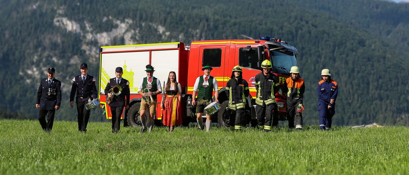 Eine Gruppe von Feuerwehrleuten und Musikern steht auf einer Wiese, mit einem roten Feuerwehrauto dahinter und einer Bergkette im Hintergrund.