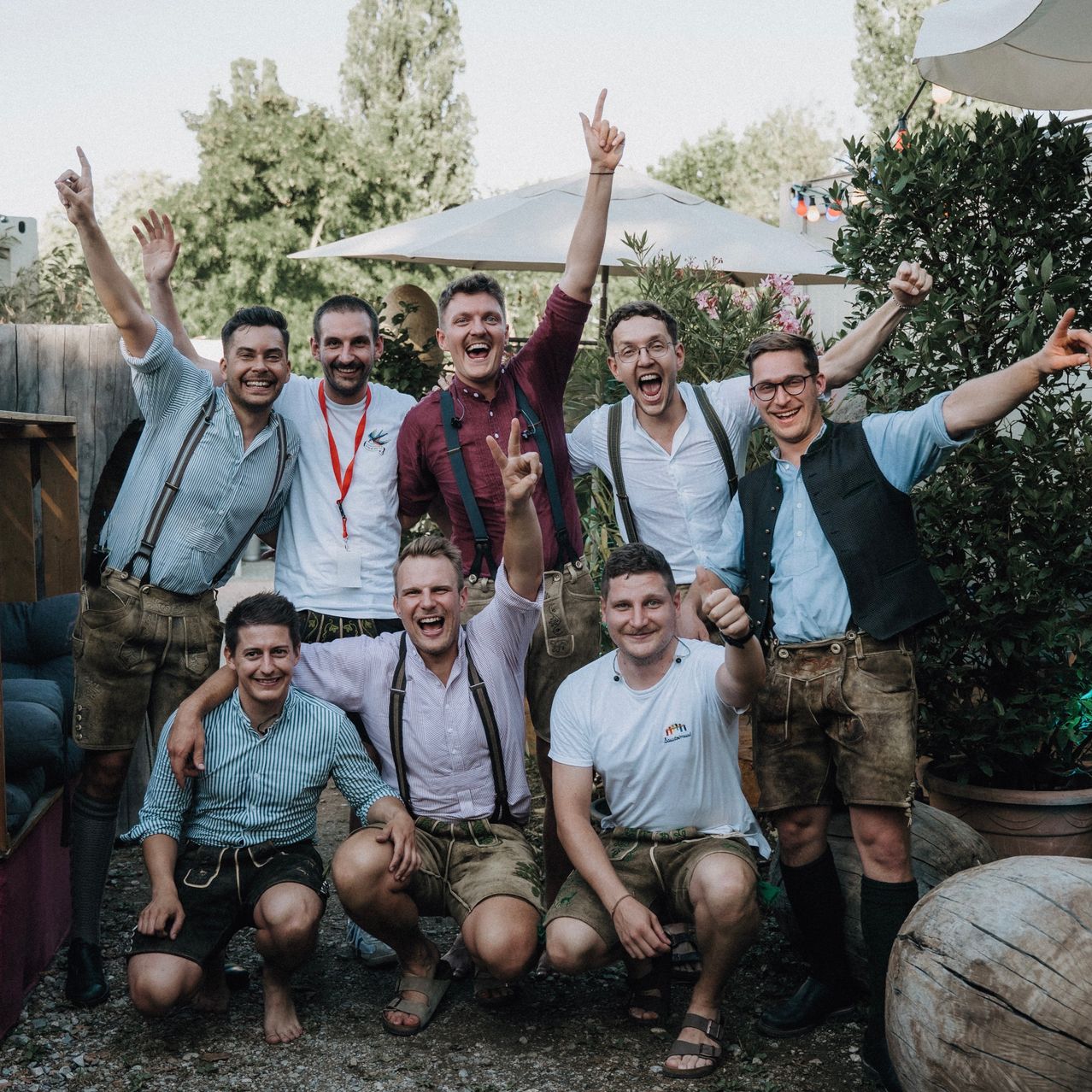 A group of men in traditional Bavarian attire pose for a photo in an outdoor garden setting, with several making humorous gestures.