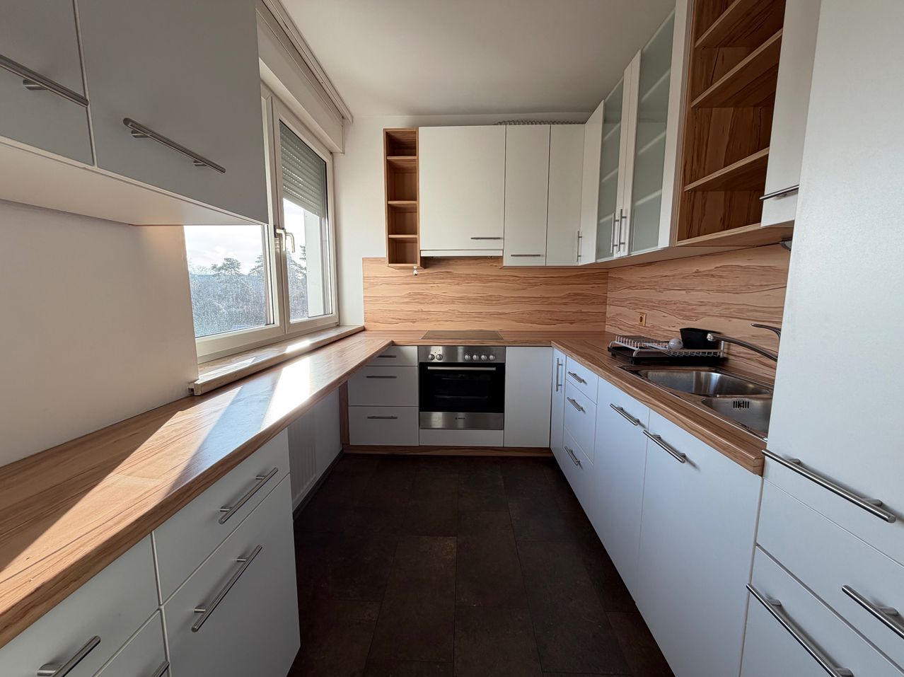 Eine leere moderne Küche mit hölzernen Arbeitsplatten, weißen Schränken, einem Edelstahlofen und einem Waschbecken mit Wasserhahn. Ein Fenster befindet sich links.