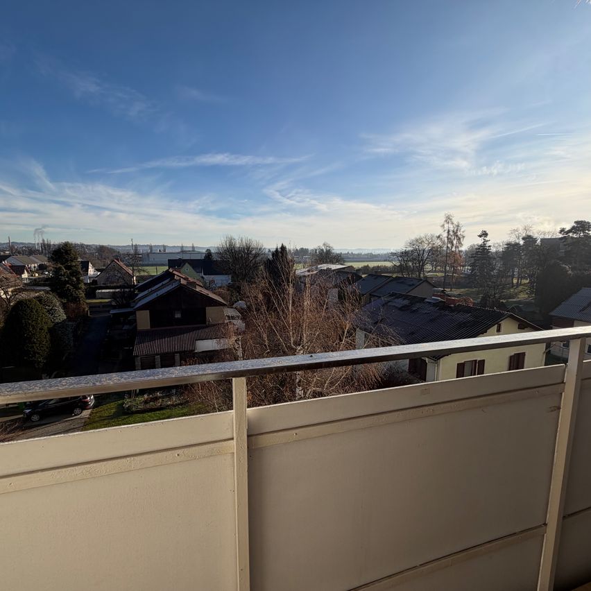 Ein Balkonblick zeigt einen klaren blauen Himmel mit einigen Wolken, ein Wohngebiet mit Häusern und in der Ferne Bäume und Gebäude.
