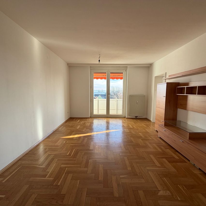 Ein leerer Raum mit Holzboden, weißen Wänden und Glastüren, die zu einem Balkon führen. Ein Holzschrank mit Schubladen und Regalen steht an der Wand.
