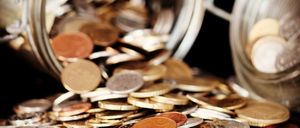 Ein Glasglas mit verschiedenen Münzen, darunter Silber-, Gold- und Kupfermünzen, ist auf einer dunklen Oberfläche umgekippt.