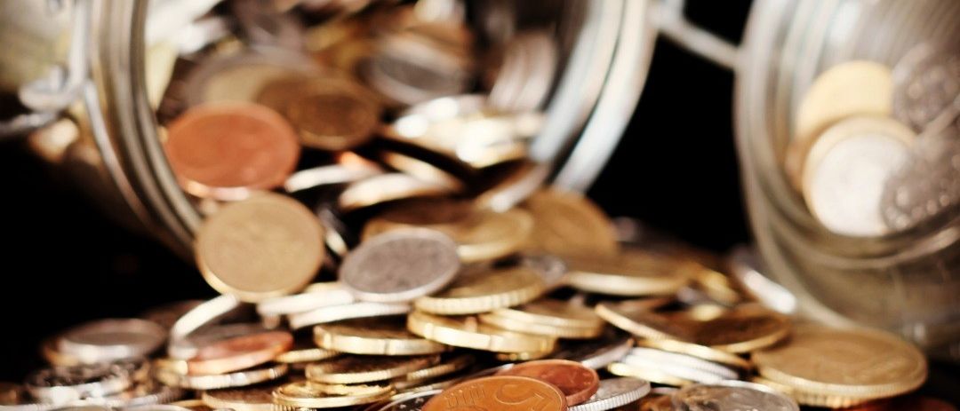 Ein Glasglas mit verschiedenen Münzen, darunter Silber-, Gold- und Kupfermünzen, ist auf einer dunklen Oberfläche umgekippt.