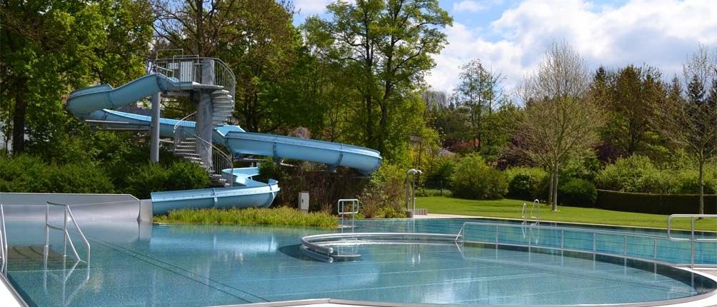 Ein Außenpool mit einer großen blauen Wasserrutsche, umgeben von Grünflächen und Bäumen. Die Rutsche führt zum Pool, der eine Sprungbrett und eine Wendeltreppe hat.