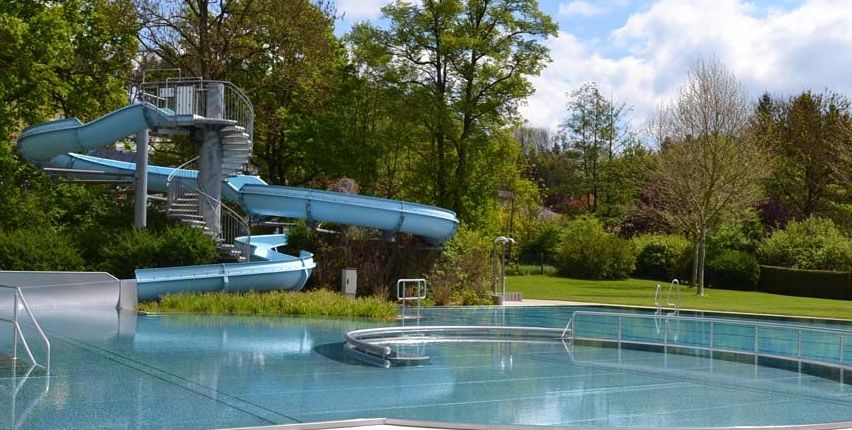 Ein Außenpool mit einer großen blauen Wasserrutsche, umgeben von Grünflächen und Bäumen. Die Rutsche führt zum Pool, der eine Sprungbrett und eine Wendeltreppe hat.