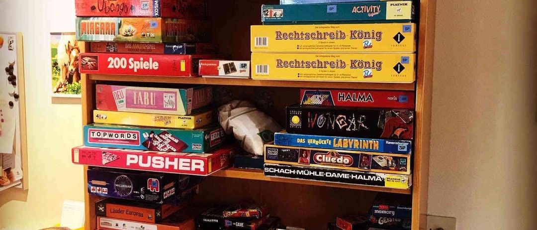 Ein hölzerne Bücherregal gefüllt mit verschiedenen Brettspielen, darunter Titel wie '200 Spiele', 'Rechschreib König' und 'PUSHER'. Die Sammlung umfasst bunte Schachteln und Puzzles, wobei einige Spiele übereinander gestapelt sind.