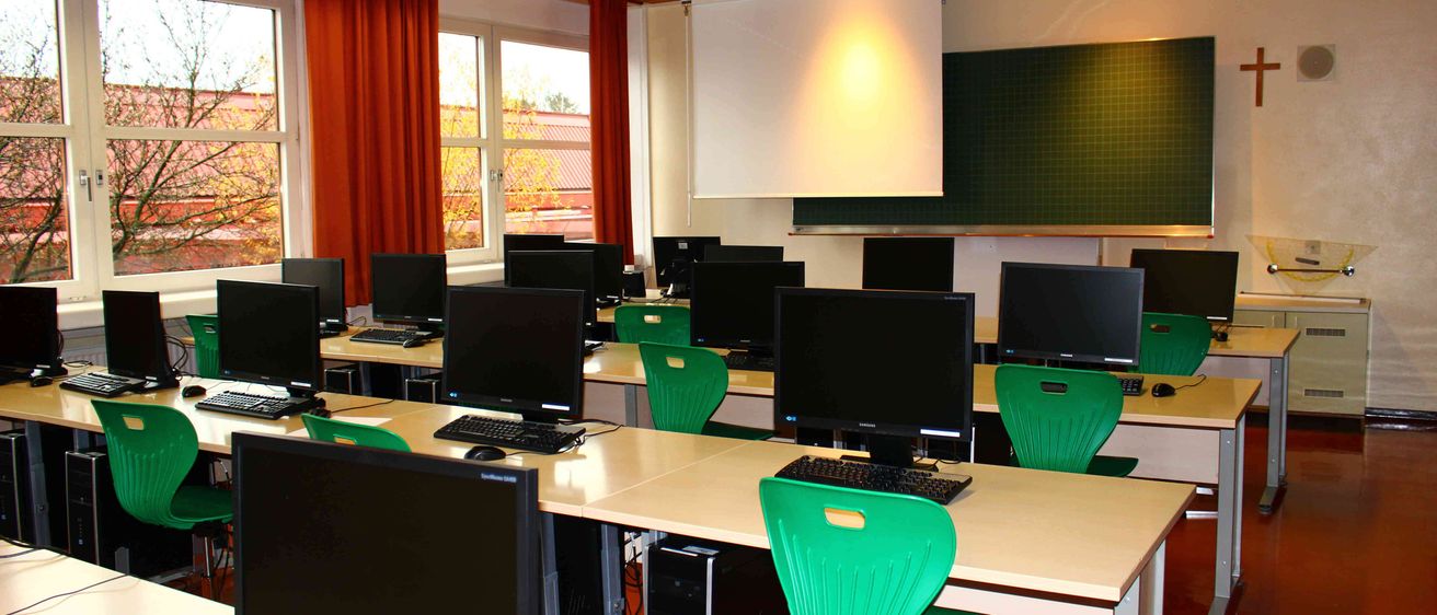 Ein Klassenzimmer mit mehreren Computern, grünen Stühlen und einem Beamer. Der Raum hat eine grüne Gittertafel und eine weiße Projektionsfläche.