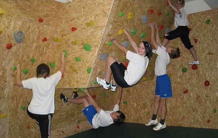Fünf Kinder klettern an einer Holzkletterwand. Einige stehen, andere hängen und einer ist kopfüber.