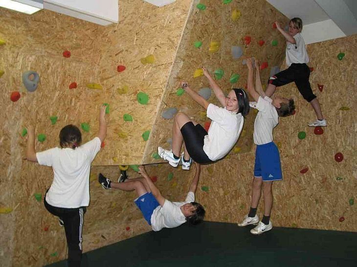 Fünf Kinder klettern an einer Holzkletterwand. Einige stehen, andere hängen und einer ist kopfüber.