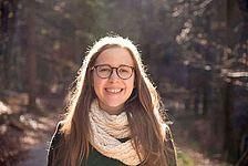 Eine Frau mit langen Haaren und Brille steht auf einem Waldweg und lächelt. Sie trägt einen weißen Schal und ein grünes Oberteil. Hinter ihr fällt Sonnenlicht durch die Bäume.