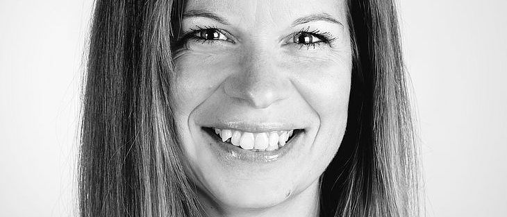 Ein schwarz-weißes Porträt einer Frau mit langen Haaren, die lächelt und nach vorne schaut. Sie hat einen Muttermal auf ihrer linken Wange.