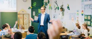 Ein Mann in einem blauen Anzug unterrichtet eine Klasse. Die Schüler sitzen an ihren Tischen, einige mit erhobenen Händen. Eine Uhr und Pflanzen schmücken die Wände.