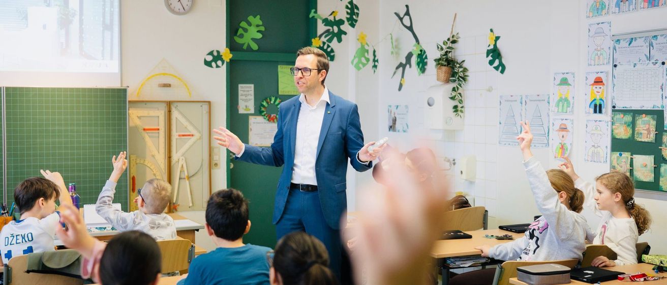 Ein Mann in einem blauen Anzug unterrichtet eine Klasse. Die Schüler sitzen an ihren Tischen, einige mit erhobenen Händen. Eine Uhr und Pflanzen schmücken die Wände.