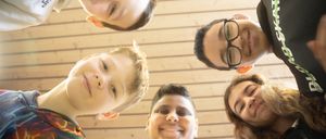 Eine Gruppe lächelnder Kinder und Jugendlicher steht im Kreis, möglicherweise für ein Foto. Die hölzerne Wand im Hintergrund verleiht der Szene eine warme Note.