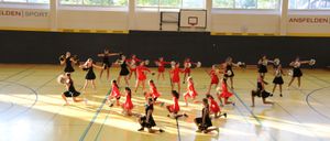 Eine Gruppe Cheerleader in roten Uniformen führt eine Routine auf einem Basketballplatz mit einem Spielstand im Hintergrund durch.