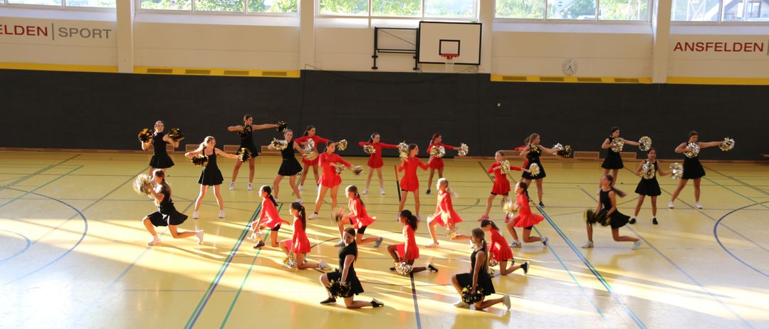Eine Gruppe Cheerleader in roten Uniformen führt eine Routine auf einem Basketballplatz mit einem Spielstand im Hintergrund durch.