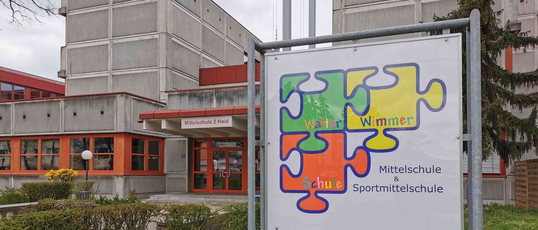 Ein Schild für Mittelschule und Sportmittelschule Walter Wimmer steht vor einem großen Gebäude mit rotem Rand und mehreren Fenstern.