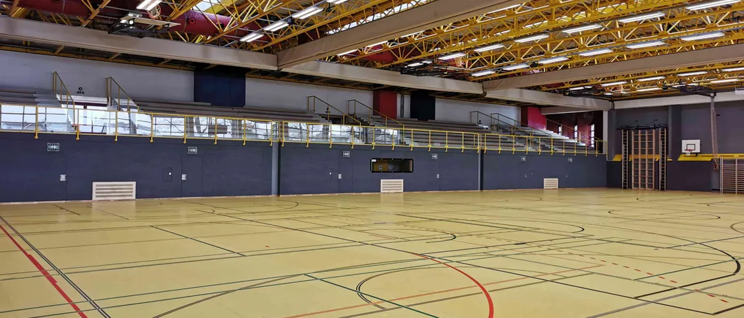Eine leere Sporthalle mit gelben Bodenmarkierungen, leeren Tribünen und gelben Stützen, die die Decke tragen.