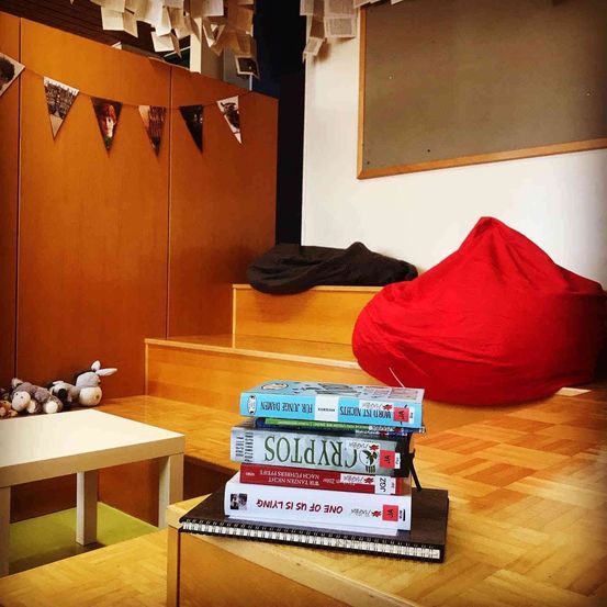 Ein Raum mit Holzregalen und einem Holzboden. Auf dem Tisch liegt ein Stapel Bücher und ein Notizbuch. Ein roter Sitzsack steht an der Wand, mit einer schwarzen Decke darauf.