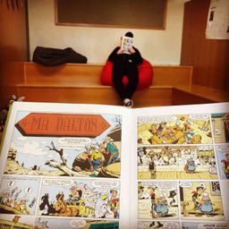 Eine Person sitzt auf einem roten Sitzsack und liest ein Comicbuch mit dem Titel Ma Dalton. Die Person trägt eine schwarze Jacke und Hose. Hinter der Person befindet sich eine Holzbank mit einer schwarzen Tasche darauf. An der Wand befindet sich eine Tafel.