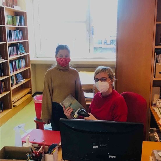 Zwei Frauen mit Gesichtsmasken in einer Bibliothek. Eine hält ein Buch, die andere sitzt an einem Computer.