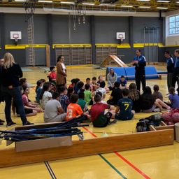 Eine Gruppe von Kindern und Erwachsenen versammelt sich in einer Sporthalle. Ein Mann steht vorne und spricht zu der Gruppe. Verschiedene Sportgeräte sind auf dem Boden sichtbar.