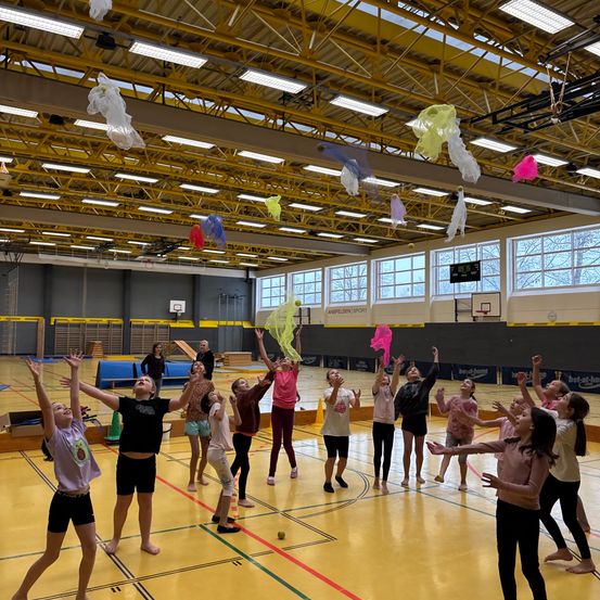 Eine Gruppe junger Mädchen übt eine Tanzroutine in einer Turnhalle, wobei sie bunte Netze halten. Sie sind von gelben Markierungen auf dem Boden und großen Fenstern umgeben.