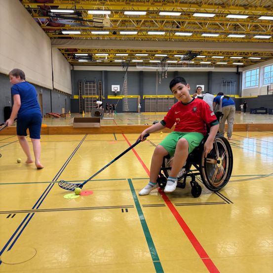 Ein junger Junge im Rollstuhl spielt ein Lacrosse-Spiel auf einem Innenplatz mit mehreren Personen um ihn herum.