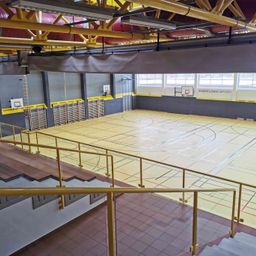 Eine Indoor-Sportanlage mit gelbem Boden und Basketballkörben an den Wänden. Gelbe Geländer und Stufen befinden sich entlang der Seite. Der Name Ansfelden Sport ist über den Körben sichtbar.