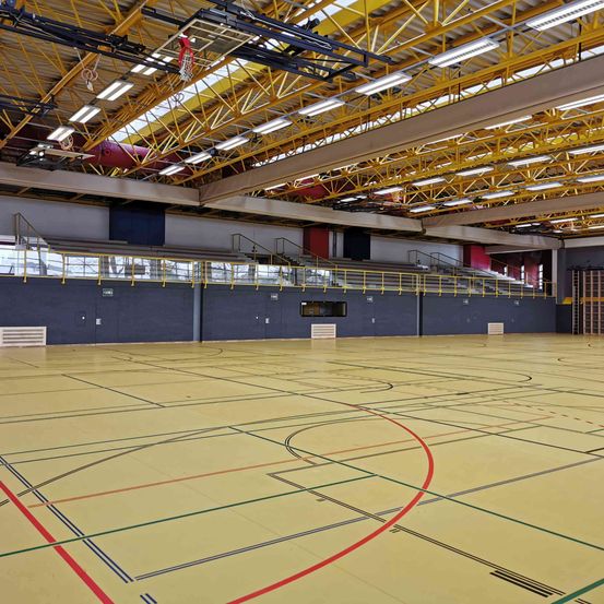 Eine leere Indoor-Basketballhalle mit gelbem Boden und roten, grünen und blauen Linien. Die Halle ist von leeren Tribünen umgeben, und die Decke wird von gelben Trägern gestützt.