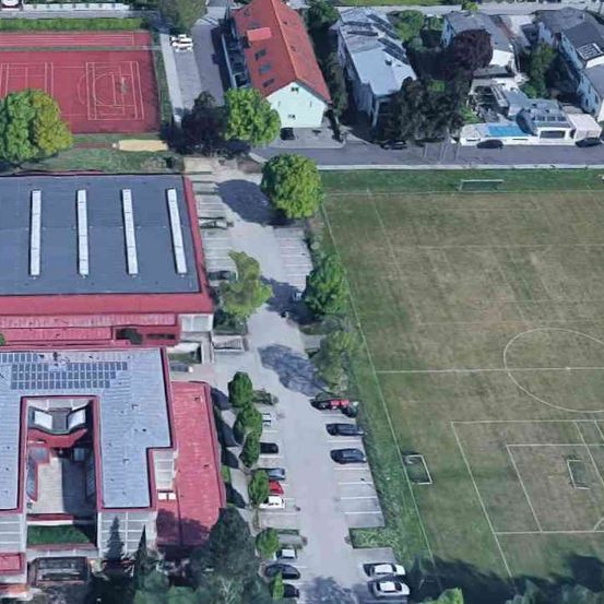 Ein Luftbild einer roten Gebäudes mit Solarpaneelen, einem Fußballfeld und einem Parkplatz mit mehreren geparkten Autos.