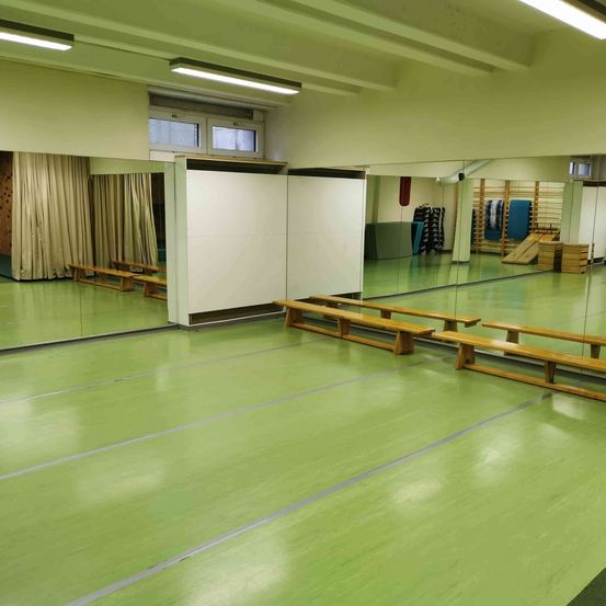 Ein leerer Raum mit grünem Boden, Holzbänken und Spiegeln. Fenster, Vorhänge und Trainingsgeräte sind sichtbar.