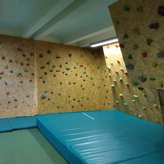 Ein Indoor-Kletterraum mit mehreren Kletterwänden, die mit farbenfrohen Griffen bedeckt sind, und einer großen blauen Schaumstoffmatte auf dem Boden.