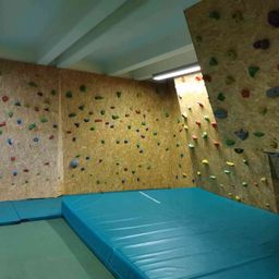 Ein Indoor-Kletterraum mit mehreren Kletterwänden, die mit farbenfrohen Griffen bedeckt sind, und einer großen blauen Schaumstoffmatte auf dem Boden.