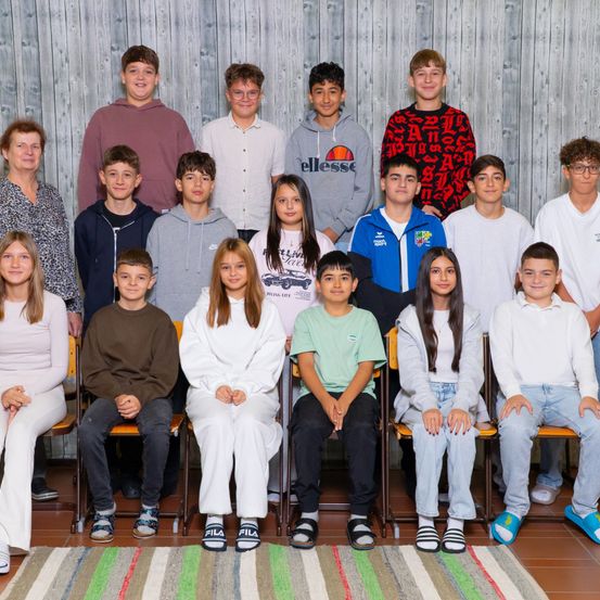 Eine Gruppe von Schülern und Lehrern posiert für ein Foto vor einer Holzwand. Einige tragen Freizeitkleidung, andere haben formelle Kleidung. Der Raum hat einen Holzboden und einen gestreiften Teppich.