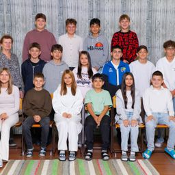 Eine Gruppe von Schülern und Lehrern posiert für ein Foto vor einer Holzwand. Einige tragen Freizeitkleidung, andere haben formelle Kleidung. Der Raum hat einen Holzboden und einen gestreiften Teppich.