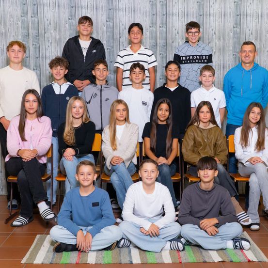 Eine Gruppe von Schülern und ein erwachsener Mann lächeln und posieren für ein Foto. Die Schüler tragen verschiedene Kleidungsstücke und sitzen auf Stühlen. Der erwachsene Mann steht hinter ihnen in einer blauen Kapuzenjacke. Die Wand hinter ihnen ist aus Holz.