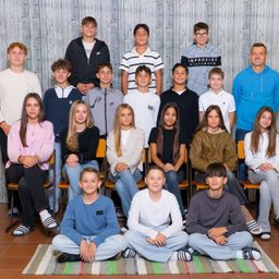 Eine Gruppe von Schülern und ein erwachsener Mann lächeln und posieren für ein Foto. Die Schüler tragen verschiedene Kleidungsstücke und sitzen auf Stühlen. Der erwachsene Mann steht hinter ihnen in einer blauen Kapuzenjacke. Die Wand hinter ihnen ist aus Holz.