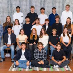 Ein Gruppenfoto von Jugendlichen in einem Klassenzimmer, einige sitzen auf Stühlen und andere auf dem Boden, vor einer Holzwand.