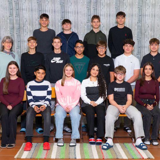 Eine Gruppe von Schülern und Lehrern posiert für ein Foto vor einem hölzernen Hintergrund. Sie sitzen auf Stühlen und einige tragen Brillen. Der Boden ist mit einem bunten Teppich bedeckt.