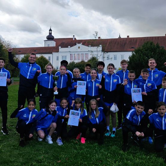 Eine Gruppe junger Athleten posiert für ein Foto auf einem Rasenfeld, alle in blauen Uniformen. Dahinter ist ein Gebäude mit einem roten Dach und weißen Wänden zu sehen, mit Bäumen und einem bewölkten Himmel im Hintergrund.