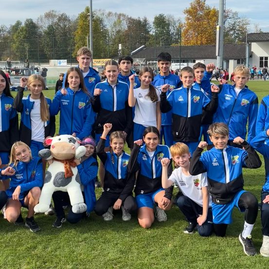 Eine Gruppe junger Athleten in blauen Uniformen posiert für ein Foto auf einem Rasenfeld mit einem Kuh-Maskottchen in der Mitte.