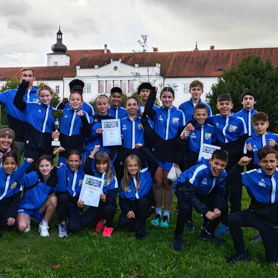 Eine Gruppe von Athleten in blauen Uniformen posiert für ein Foto vor einem großen Gebäude und hält Pokale und Urkunden in den Händen.