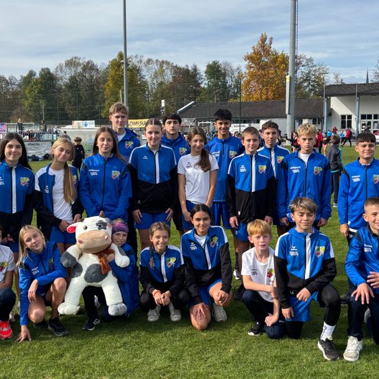 Eine Gruppe junger Athleten in blauen Uniformen posiert für ein Foto auf einem grünen Feld mit einem Kuh-Plüschtier in der Mitte. Im Hintergrund sind Bäume, ein Zaun und ein Gebäude zu sehen.