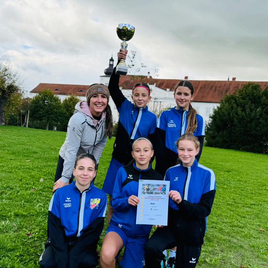 Eine Gruppe junger Athleten posiert für ein Foto auf einem Rasenfeld. Sie halten eine Trophäe und ein Zertifikat in der Hand, eine Person lächelt und schaut in die Kamera. Hinter ihnen steht ein Gebäude mit einem braunen Dach, umgeben von Bäumen.