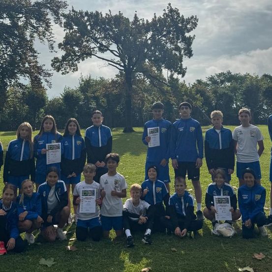 Eine Gruppe von Kindern und Jugendlichen posiert für ein Foto auf einem Rasenfeld mit Bäumen im Hintergrund. Sie tragen blaue und weiße Sportuniformen. Einige von ihnen halten Urkunden.